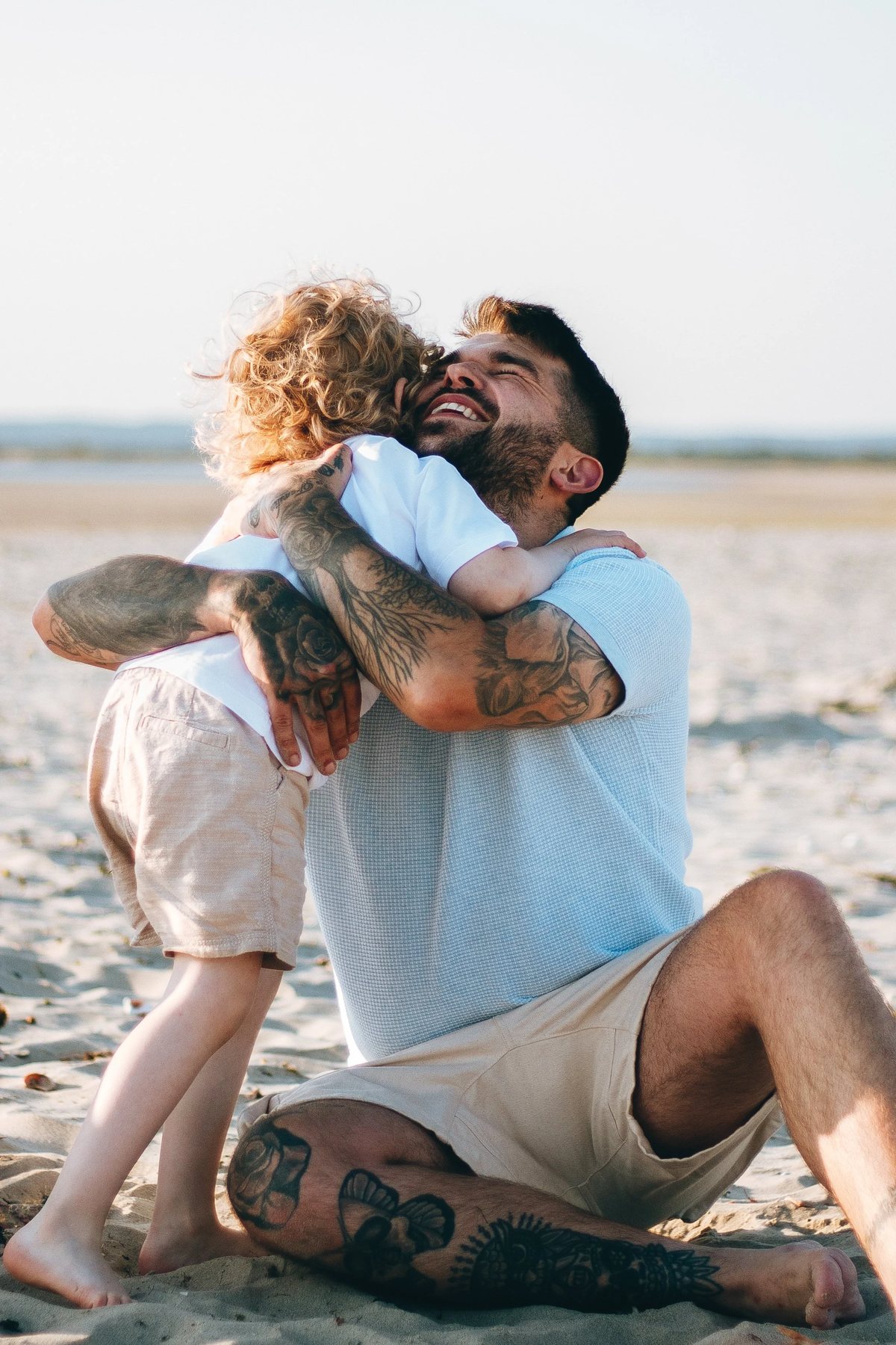 A father kneeling on wet sand lifting a small child against a pale evening sky